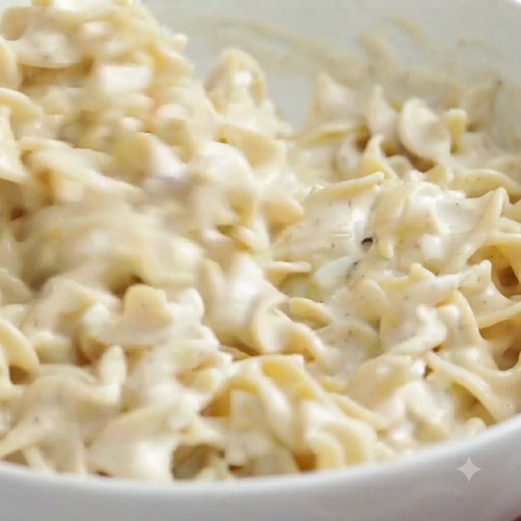 Fideos de huevo siendo revueltos en la salsa blanca cremosa usando una espátula azul de goma dentro de un tazón.