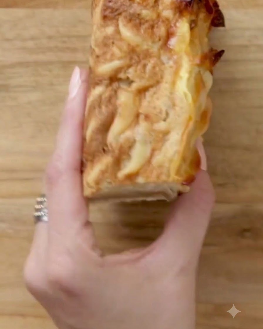 Una mano sosteniendo una porción cuadrada de kugel cortada ordenadamente sobre una tabla de madera.