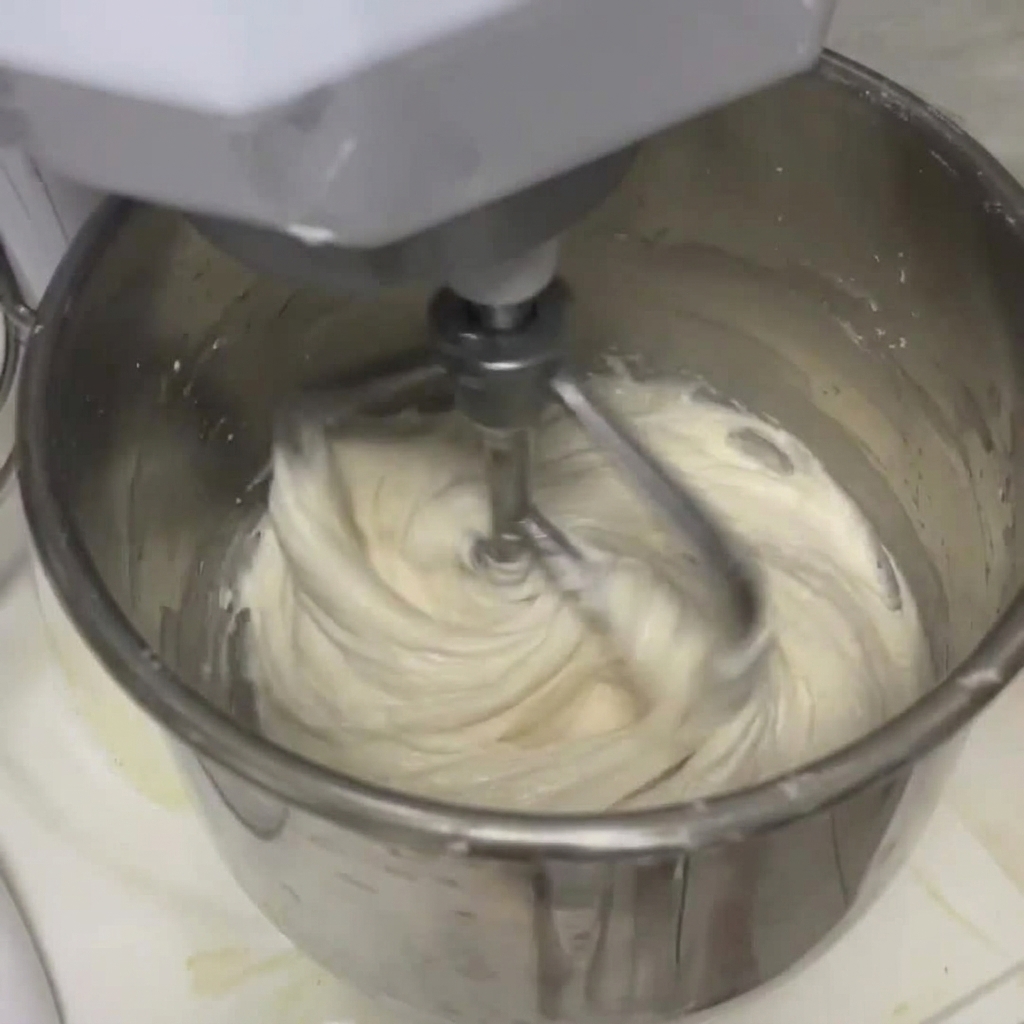 Masa blanca combinándose con líquido dentro del bol de una batidora amasadora grande utilizando un gancho para masa.