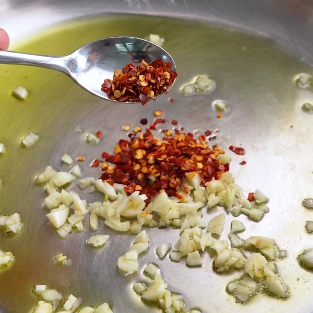 Ajo picado y hojuelas de chile rojo chisporroteando en una sartén de acero inoxidable con aceite de oliva.
