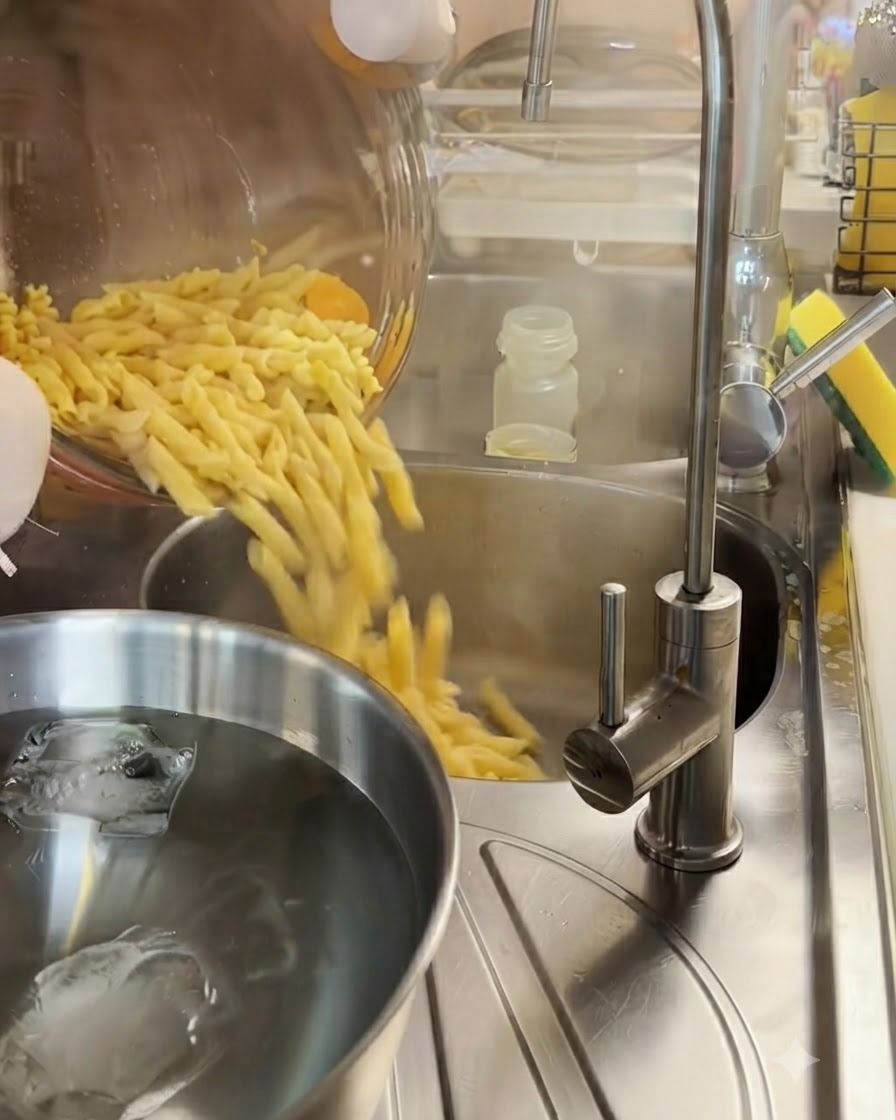 Pasta fusilli cocida escurriéndose en un colador de metal sobre un tazón de agua con hielo.