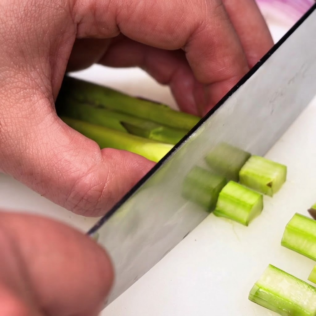 Tallos de espárragos verdes pelados siendo cortados en trozos pequeños y uniformes usando un cuchillo de chef.