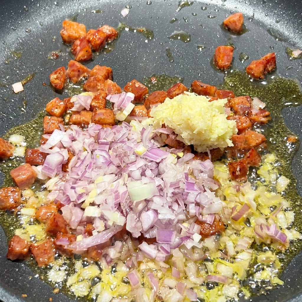 Cebolla roja en dados y ajo picado siendo agregados a la sartén caliente que contiene trozos de chorizo dorados.