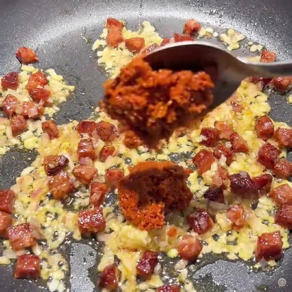Cuchara agregando pasta espesa de tomates secos y pasta de tomate a la sartén con la mezcla de chorizo y cebolla.
