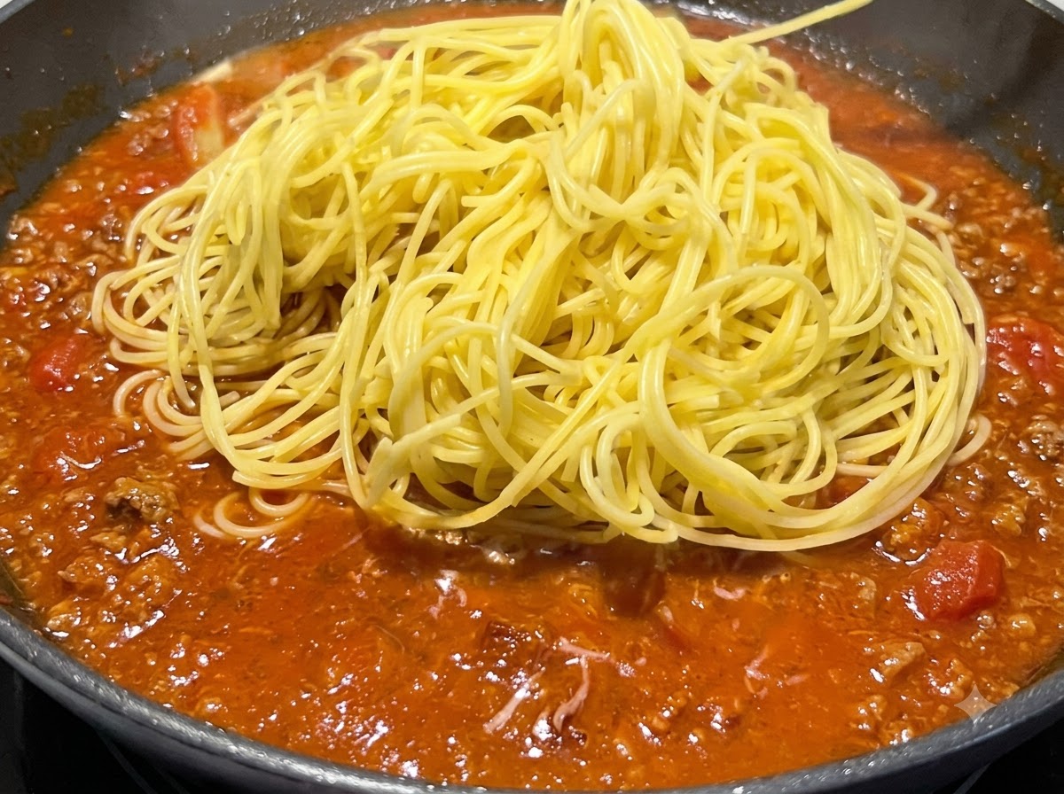 Un montón de espaguetis amarillos cocidos colocados encima de la salsa roja a fuego lento en una sartén.