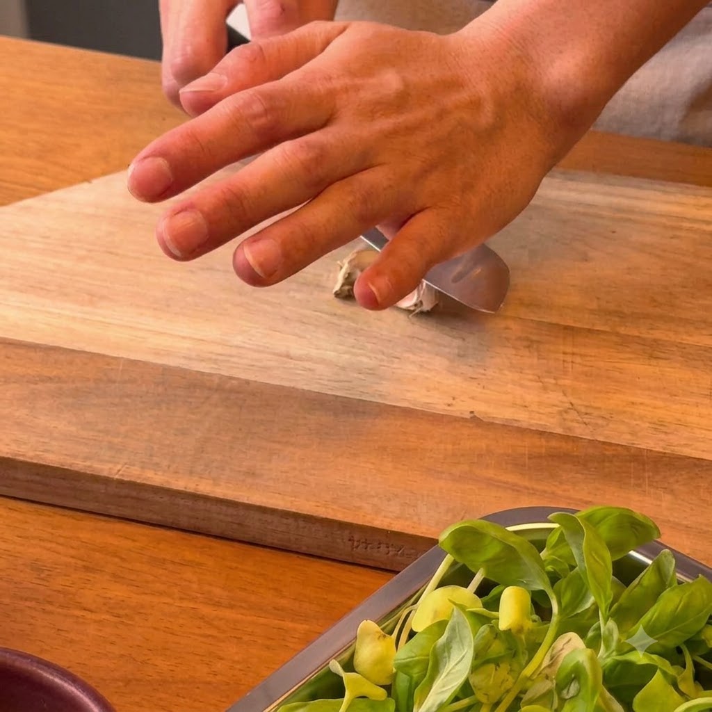 Una mano presionando el lado plano de un cuchillo de chef hacia abajo sobre un diente de ajo en una tabla de cortar de madera.