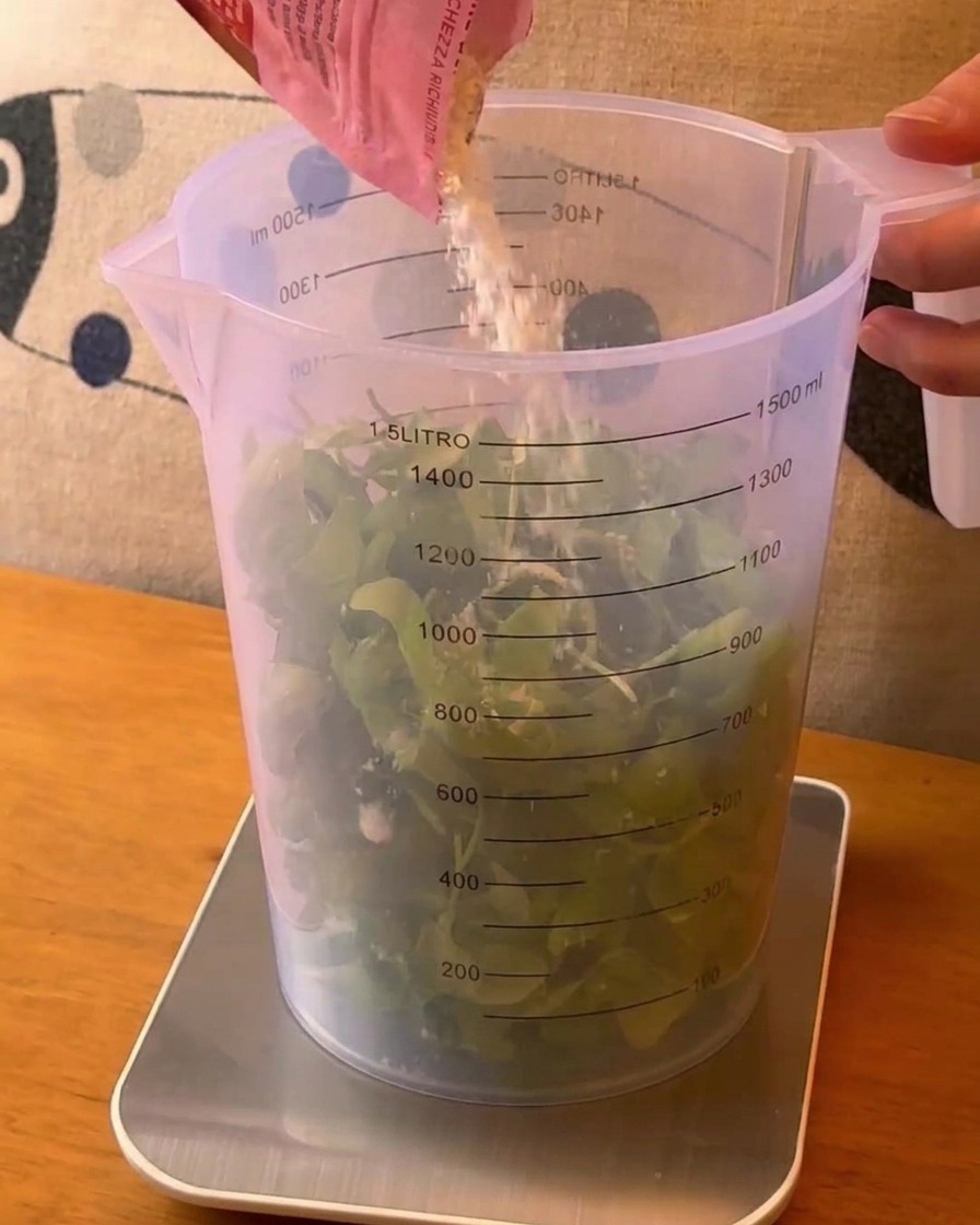 Queso rallado siendo vertido desde una bolsa rosa en un vaso medidor grande lleno de hojas de albahaca verde fresca.