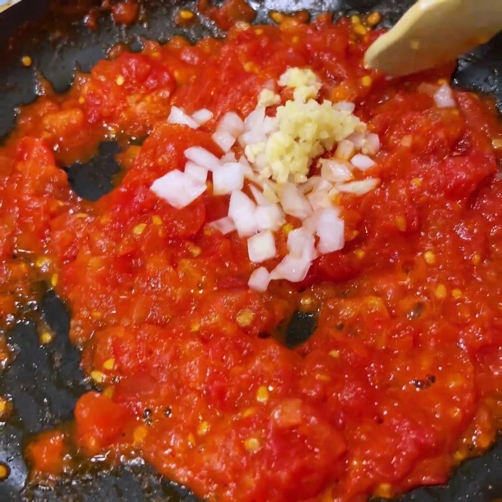 Ajo picado y cebolla blanca mezclándose con tomates triturados ablandados en una sartén.