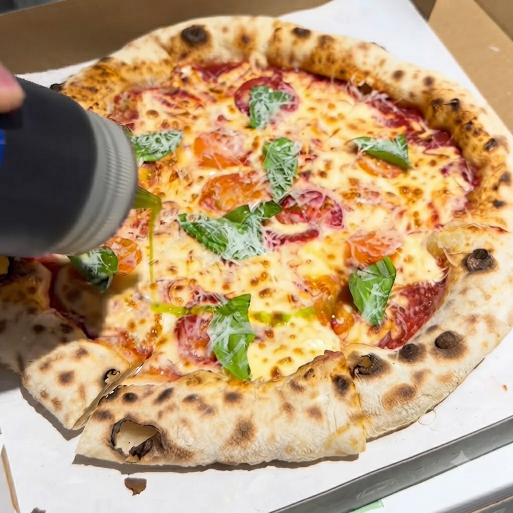 Una botella vertiendo un chorro de aceite de oliva virgen extra sobre una pizza recien horneada cubierta con albahaca y queso dentro de una caja.