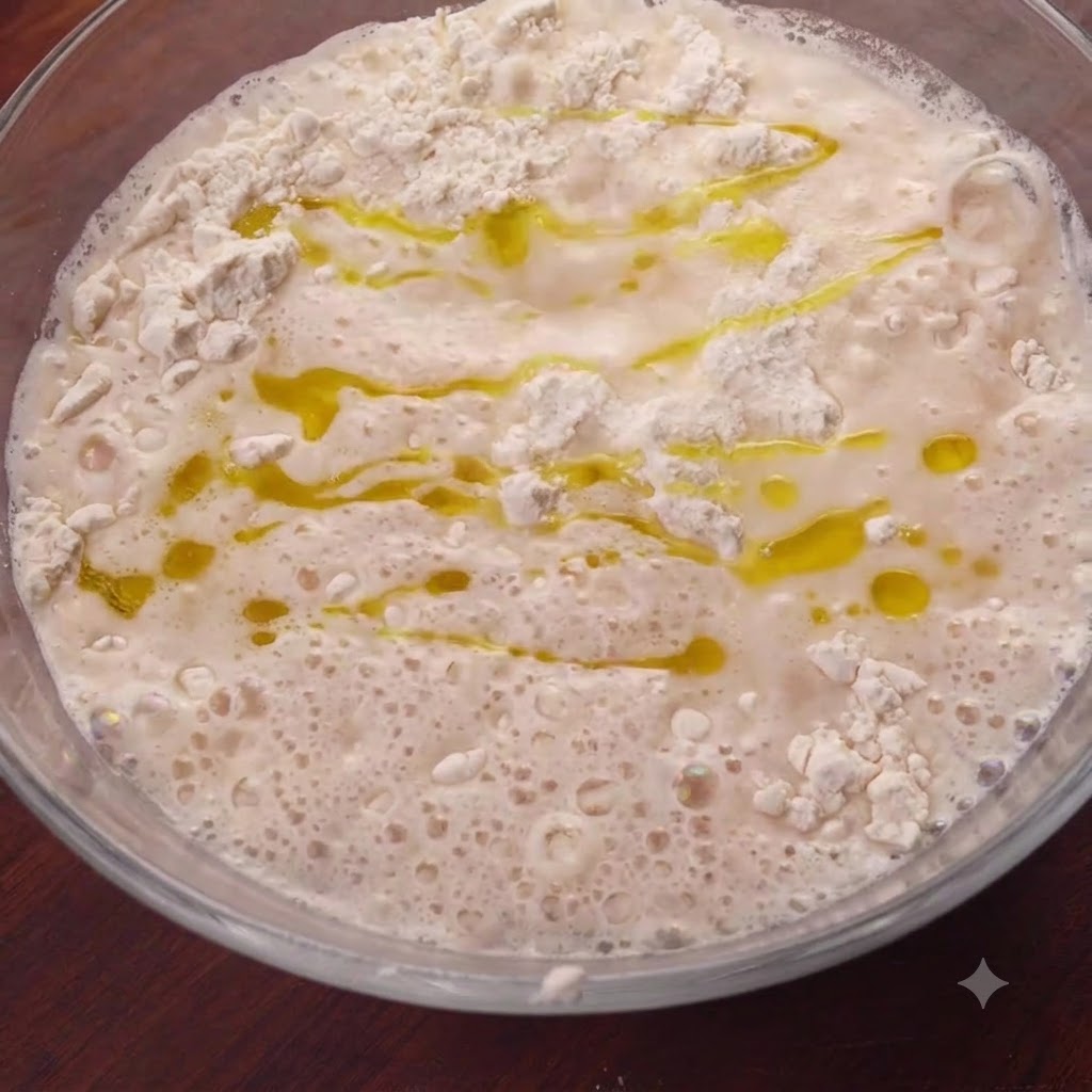 Aceite de oliva dorado rociado sobre la superficie de la mezcla de harina húmeda dentro de un tazón de vidrio.