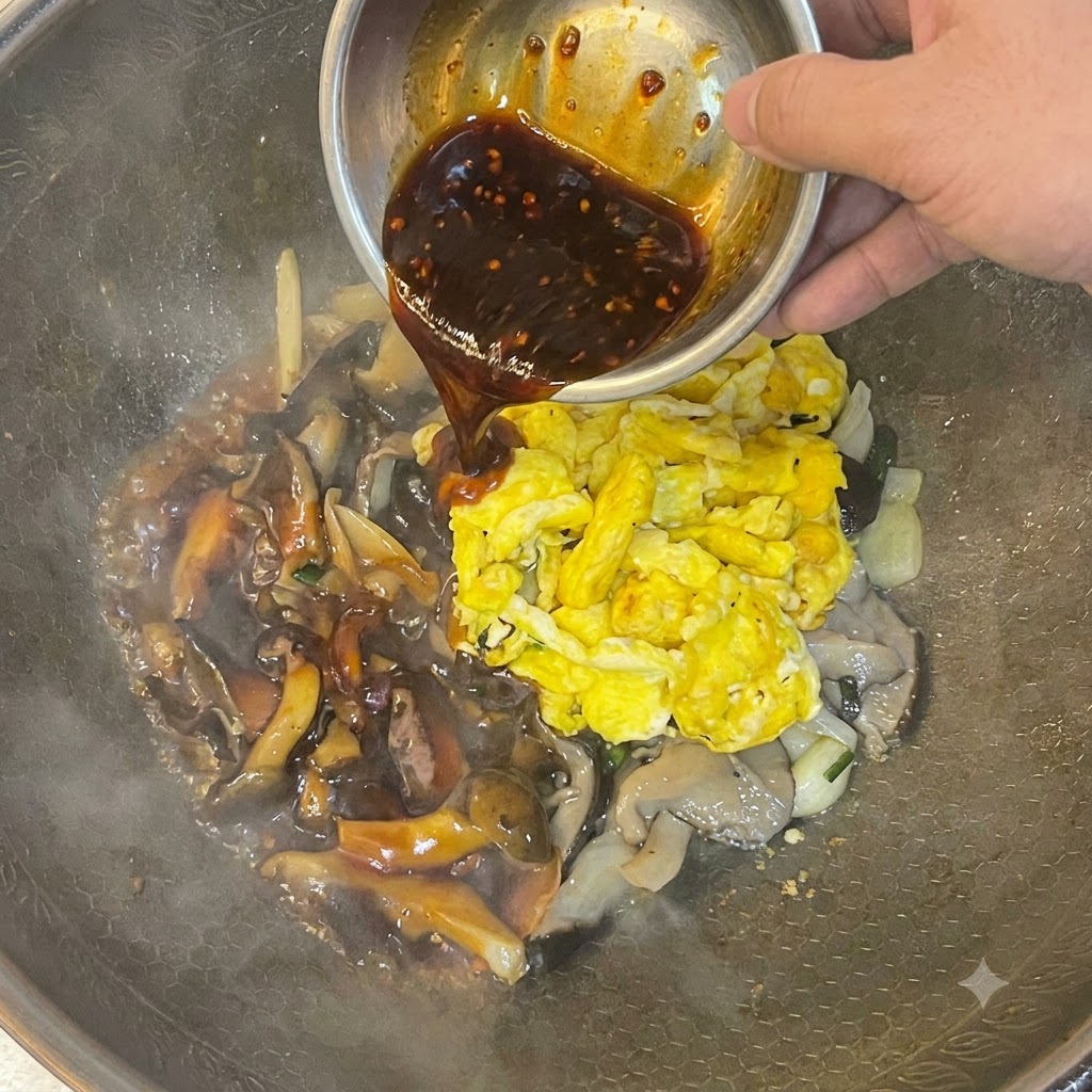 Salsa oscura siendo vertida desde un tazón de metal sobre los huevos revueltos y champiñones en un wok.