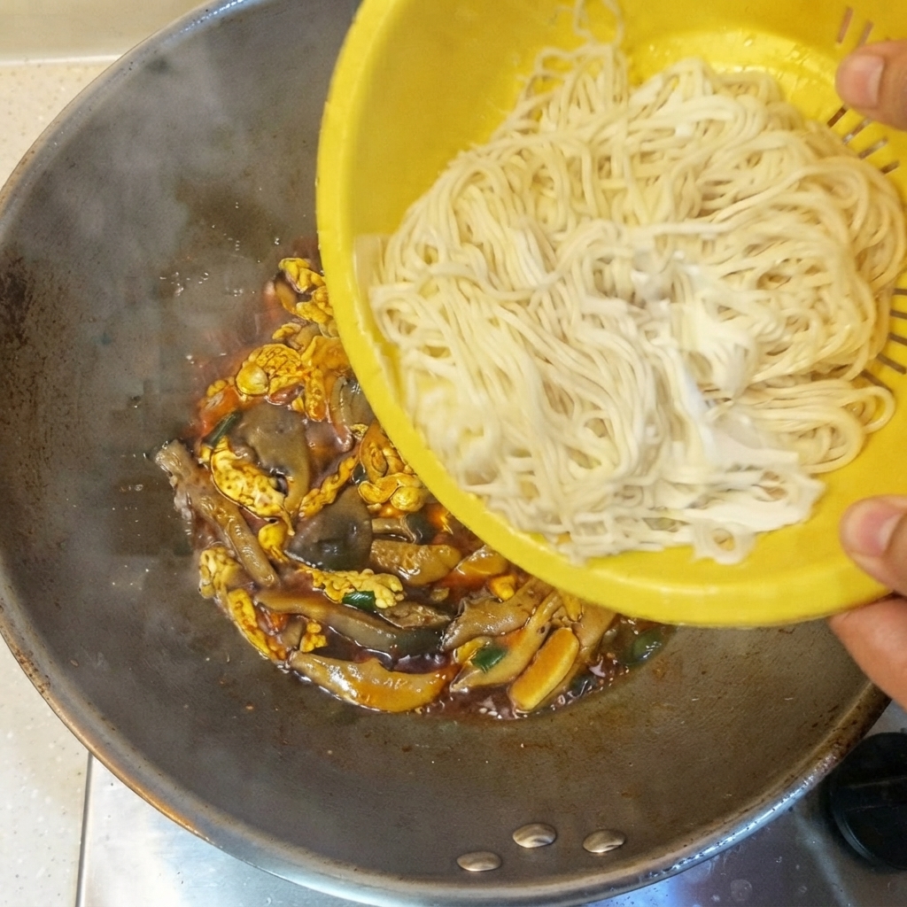 Fideos finos cocidos siendo vaciados desde un colador amarillo al wok que contiene la salsa y los ingredientes.