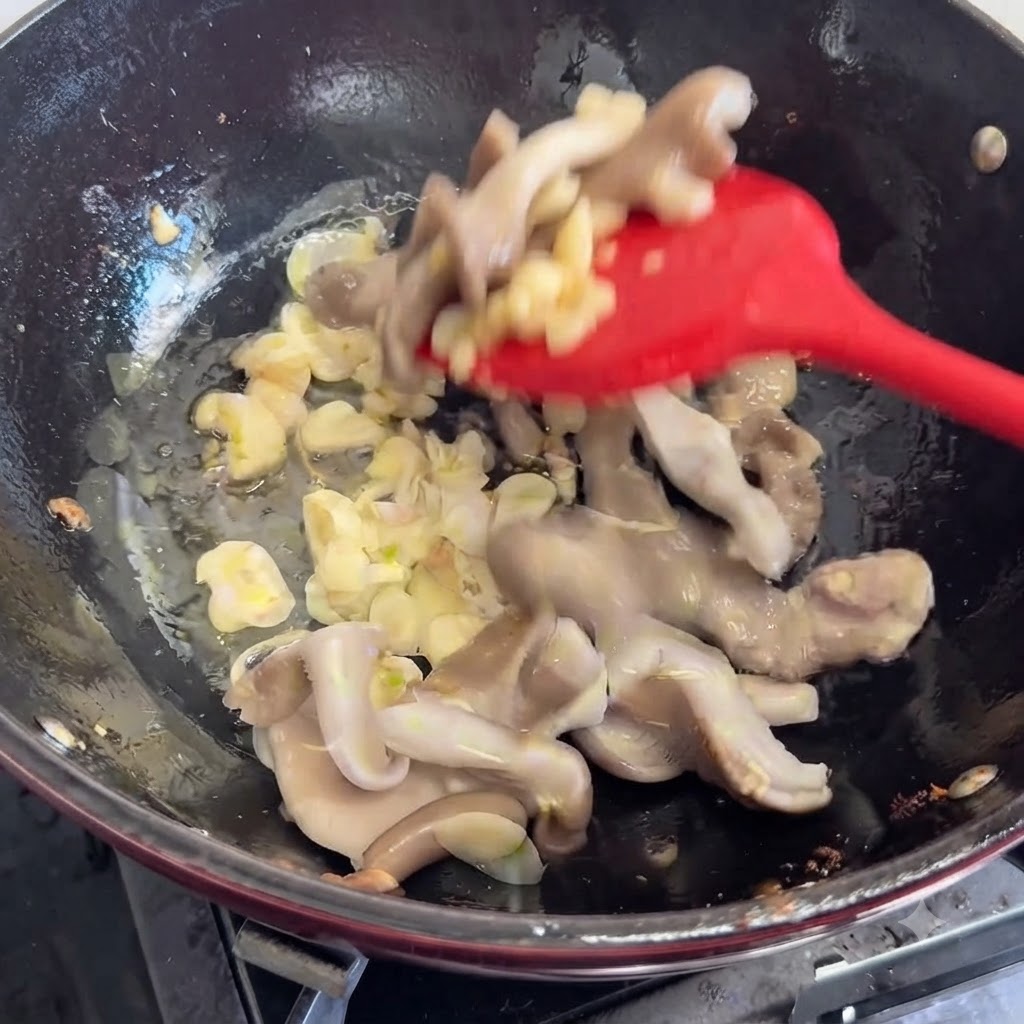 Hongos frescos siendo añadidos y revueltos en la sartén con aromáticos usando una espátula roja.