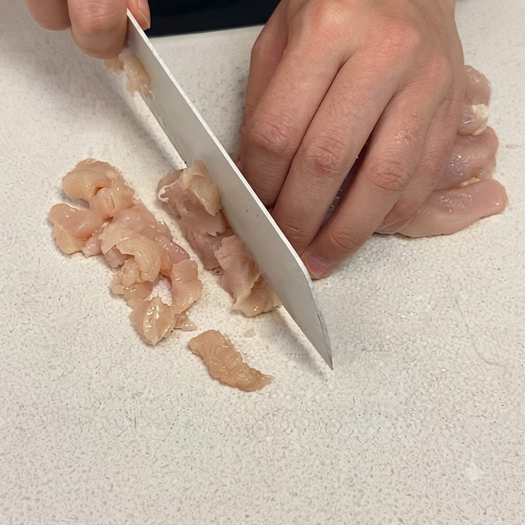 Un primer plano de una pechuga de pollo cortándose en cubos pequeños sobre una tabla de cortar blanca con un cuchillo afilado.
