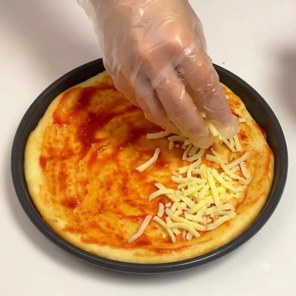 Una mano enguantada esparciendo queso mozzarella rallado sobre la base de salsa de tomate en un molde redondo para pizza.