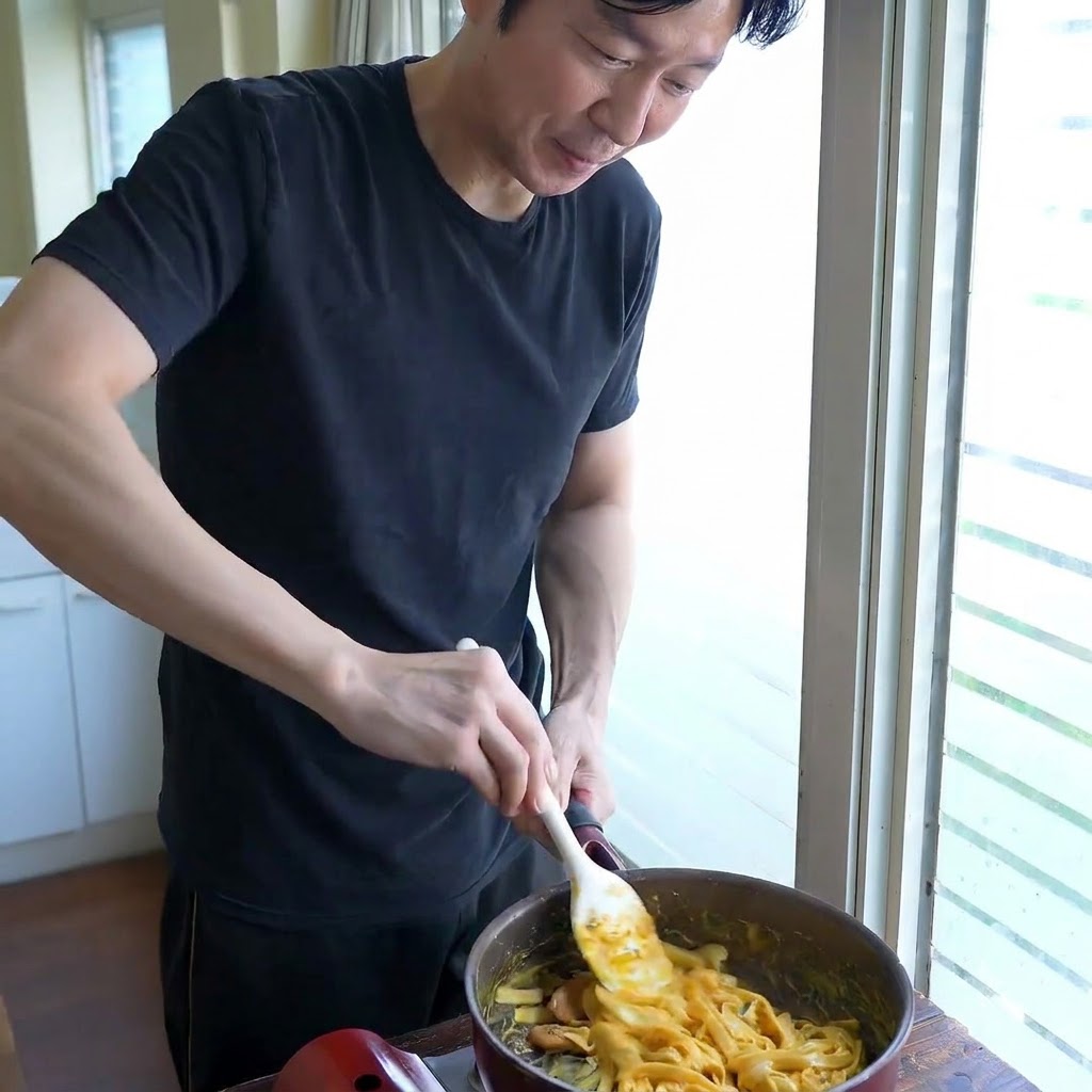 Una persona revolviendo fideos anchos en una sartén con una espátula blanca mientras ajusta la consistencia de la salsa.