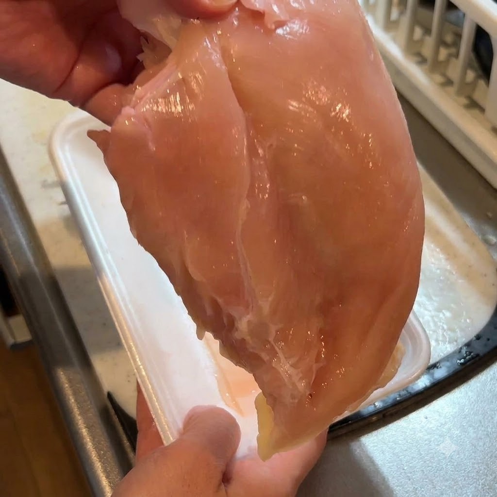Una pechuga de pollo cruda sostenida en el aire mientras se le retira la piel con un cuchillo.