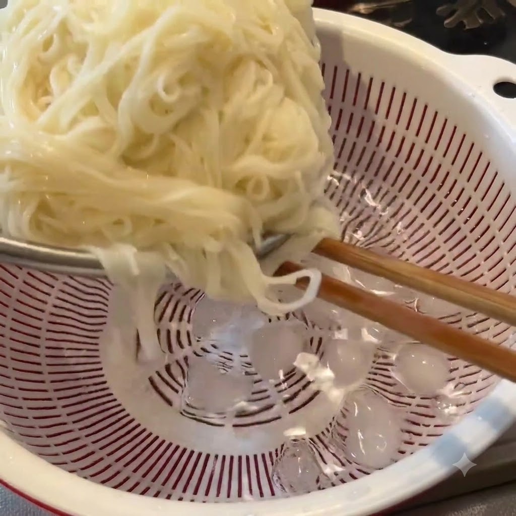 Fideos cocidos siendo enjuagados con palillos dentro de un colador blanco lleno de cubos de hielo y agua fría.