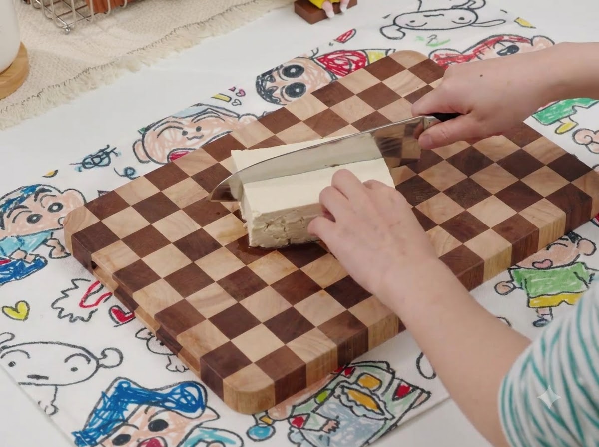 Un cuchillo cortando un bloque de tofu blanco en una tabla de cortar de madera con una estera estampada debajo.