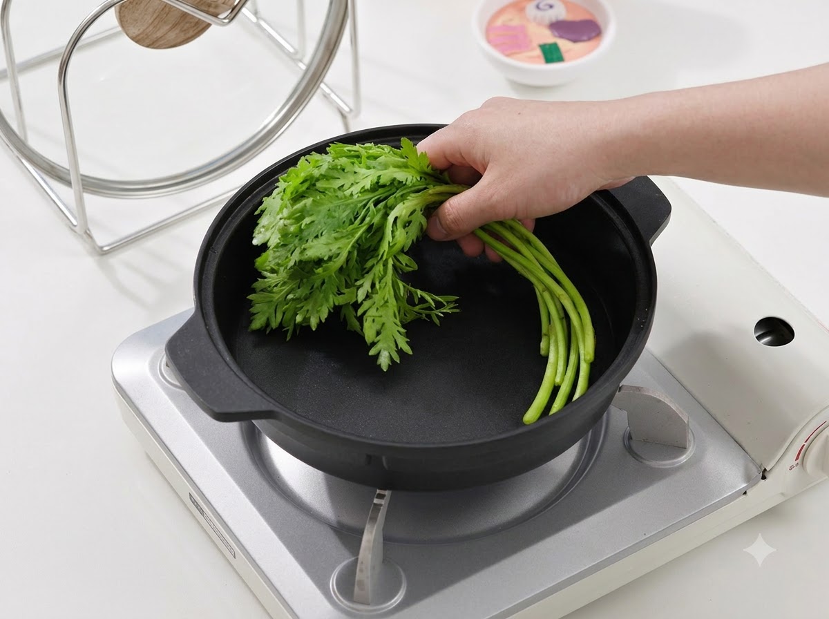 Una mano colocando un manojo de verduras de hoja verde fresca en una sartén de cocina negra sobre una estufa portátil.