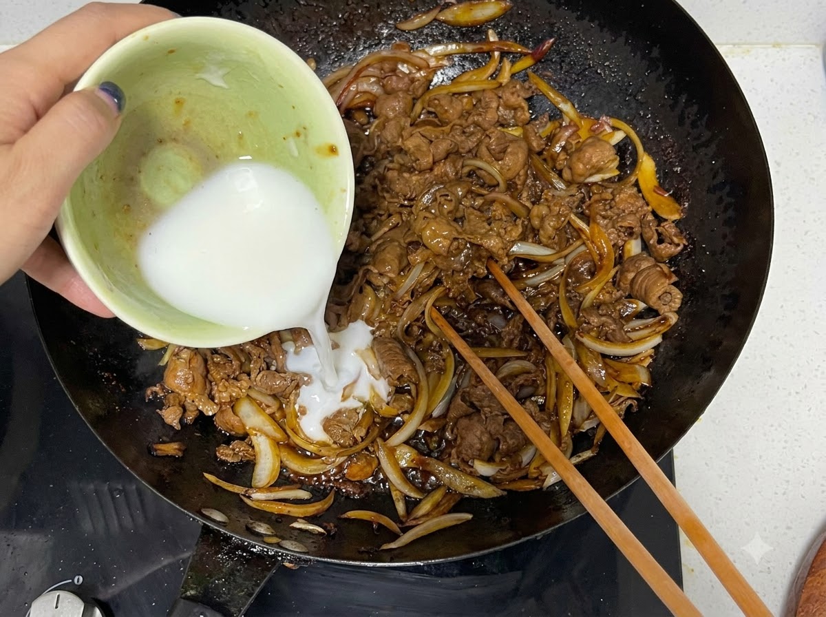 Mezcla de fécula de maíz siendo vertida desde un tazón pequeño en un wok con carne y cebollas cocidas, junto con palillos de madera.