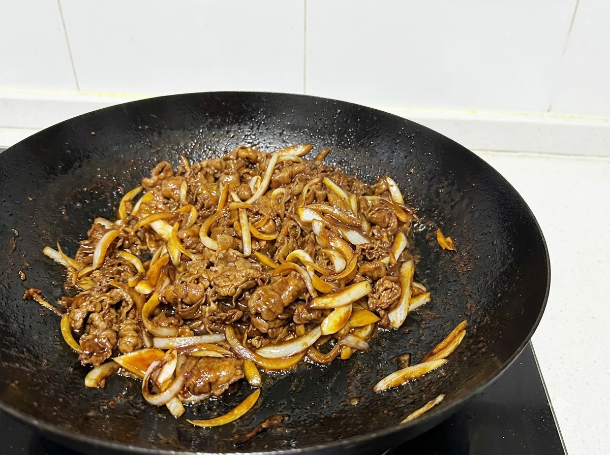 Primer plano de láminas de ternera y tiras de cebolla siendo salteadas en un wok negro con utensilios de cocina.