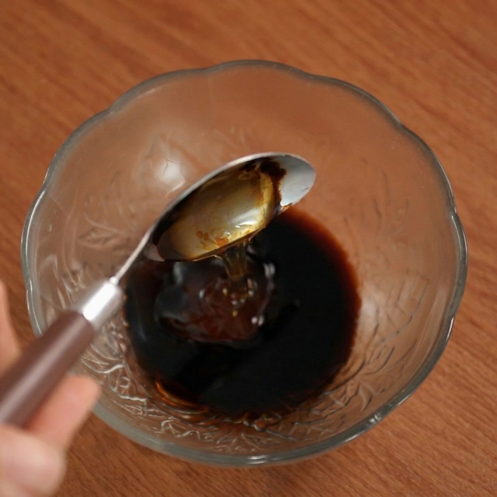 Miel siendo vertida desde una cuchara en la mezcla de salsa oscura dentro de un bol de cristal.