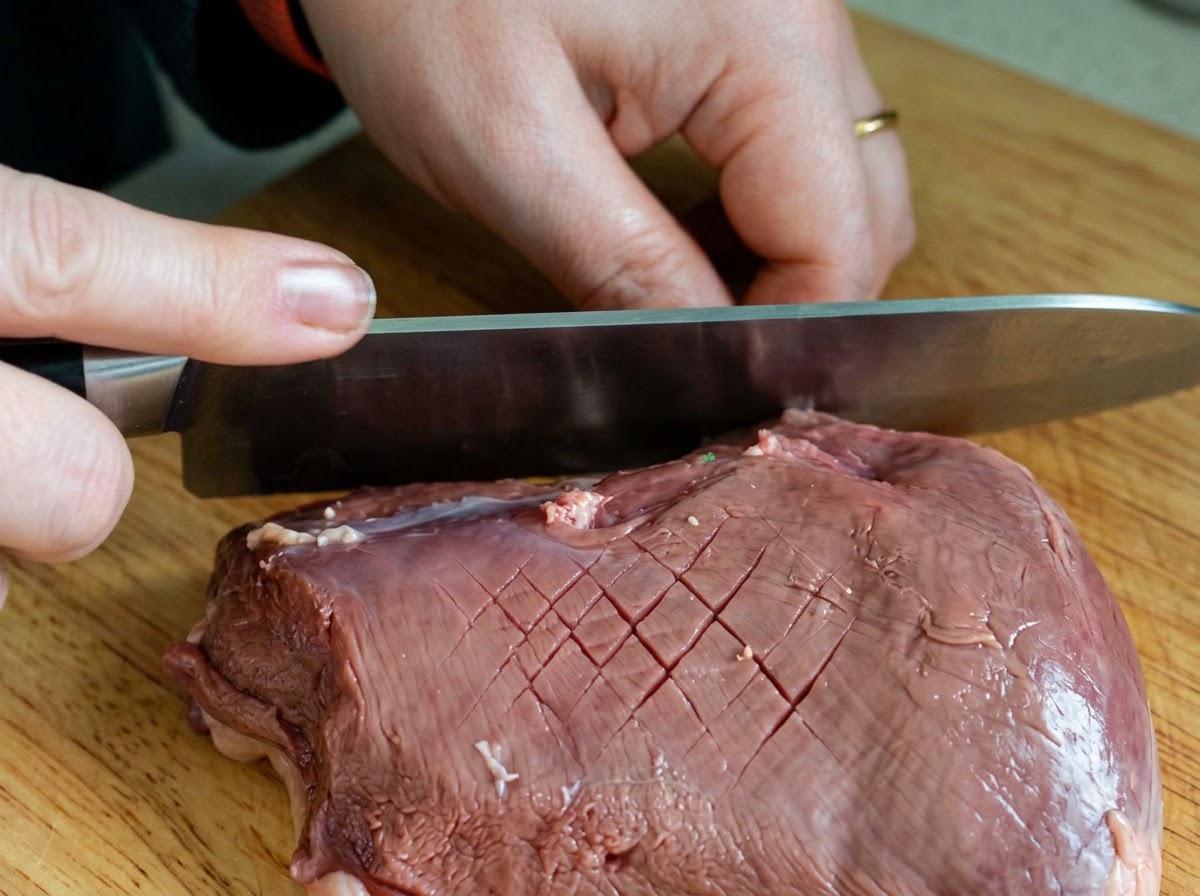Un cuchillo de chef haciendo un patrón cruzado en forma de diamante sobre la piel cruda de un pato en una tabla de madera.