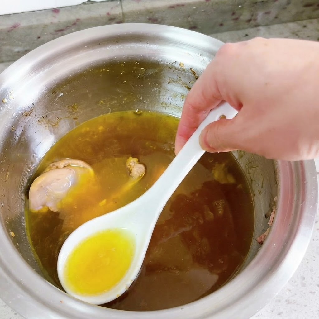 Una cuchara blanca quitando aceite amarillo brillante de la superficie de una sopa de pato a fuego lento.