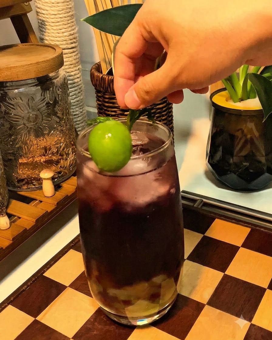 Una mano colocando una uva verde vibrante y una ramita de menta en el borde de un vaso de bebida espumosa morada.