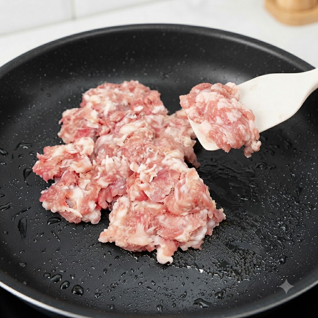 Carne de cerdo picada cruda siendo añadida y salteada en una sartén negra con una espátula de silicona.