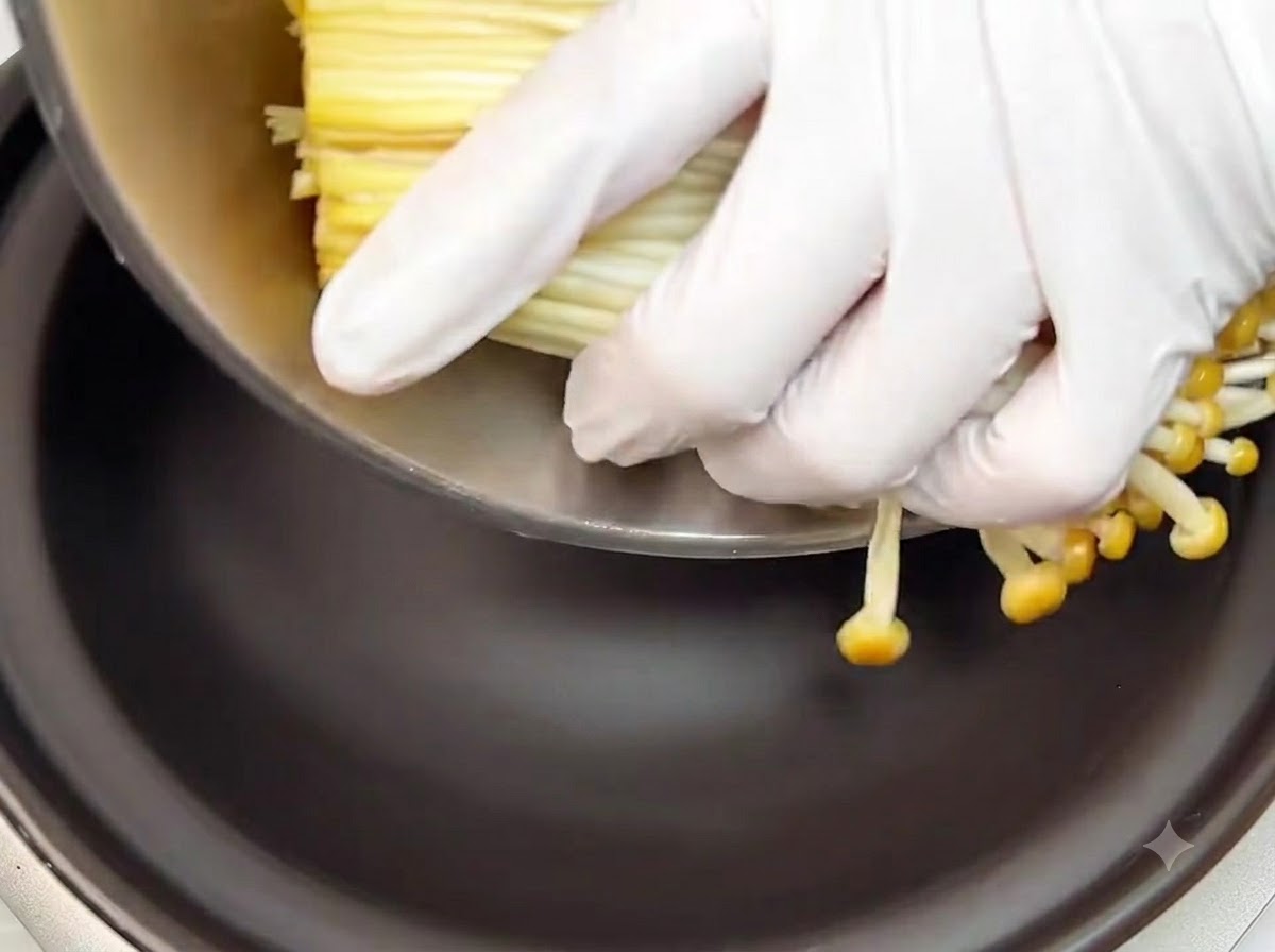 Manos con guantes colocando un manojo de champiñones enoki crudos recortados en el fondo de una olla de barro negra.