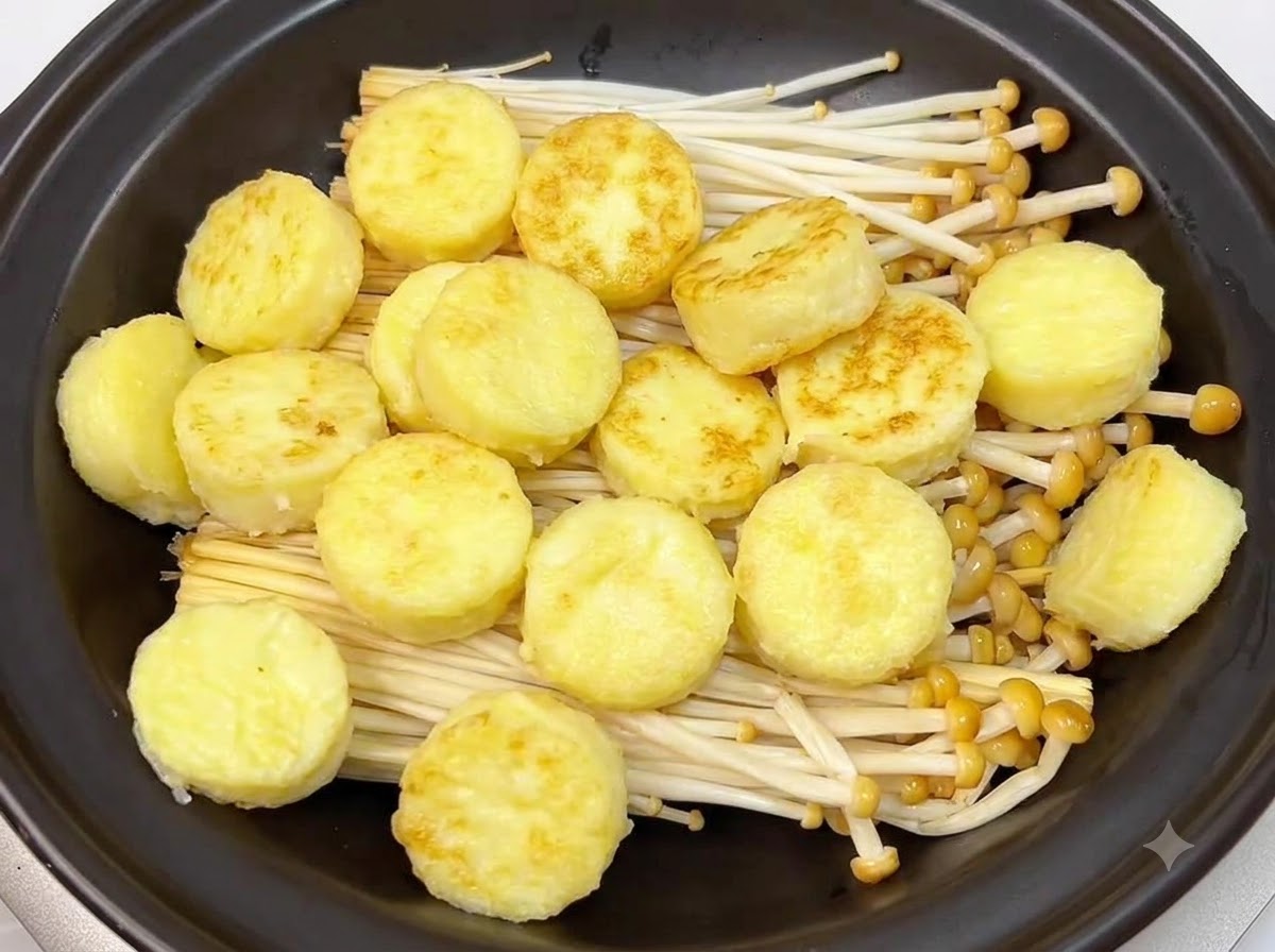 Rodajas doradas de tofu japonés fritas dispuestas ordenadamente sobre los champiñones enoki dentro de una olla de barro negra.