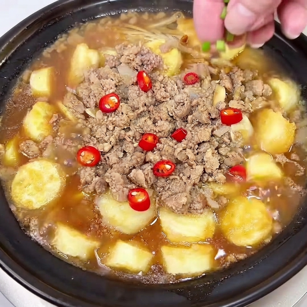 Una mano espolvoreando cebolletas picadas y rodajas de chile rojo sobre el plato de olla de barro hirviendo a fuego lento.