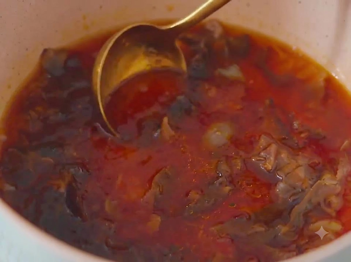 Un primer plano de una cuchara de metal dorada removiendo una mezcla de salsa de color marrón rojizo con copos de bonito dentro de una olla de colores claros.