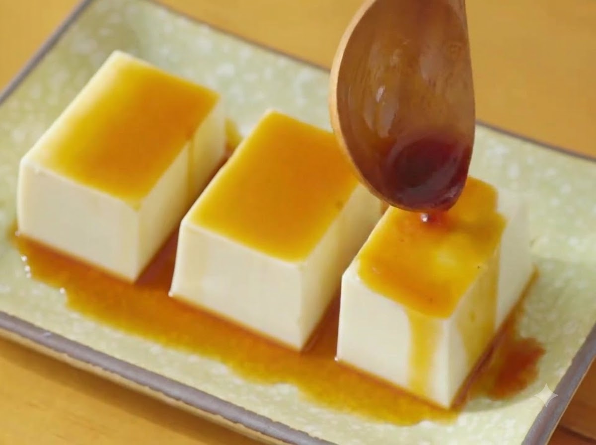 Una cuchara de madera vertiendo salsa de soja oscura sobre tres filas de tofu sedoso en un plato verde.