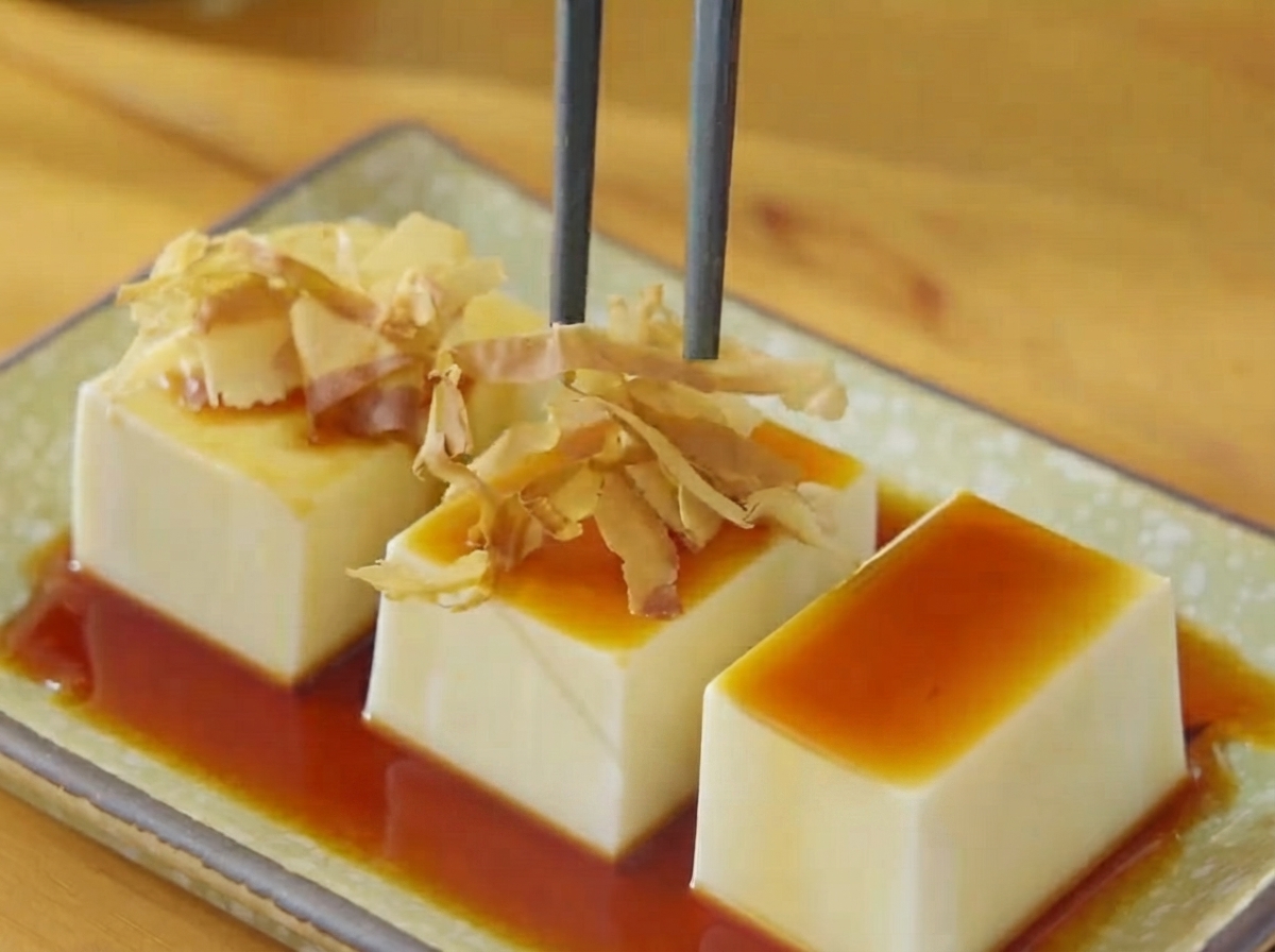 Palillos colocando copos de bonito secos y tiras de alga sobre el tofu rociado con salsa de soja.