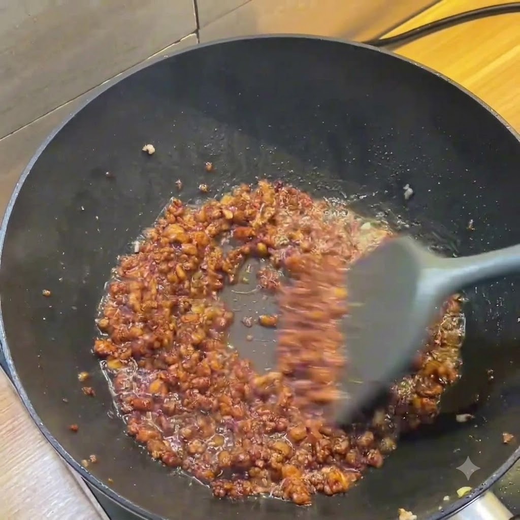 Un wok oscuro que muestra una rica salsa marrón de carne picada siendo revuelta con una espátula gris.