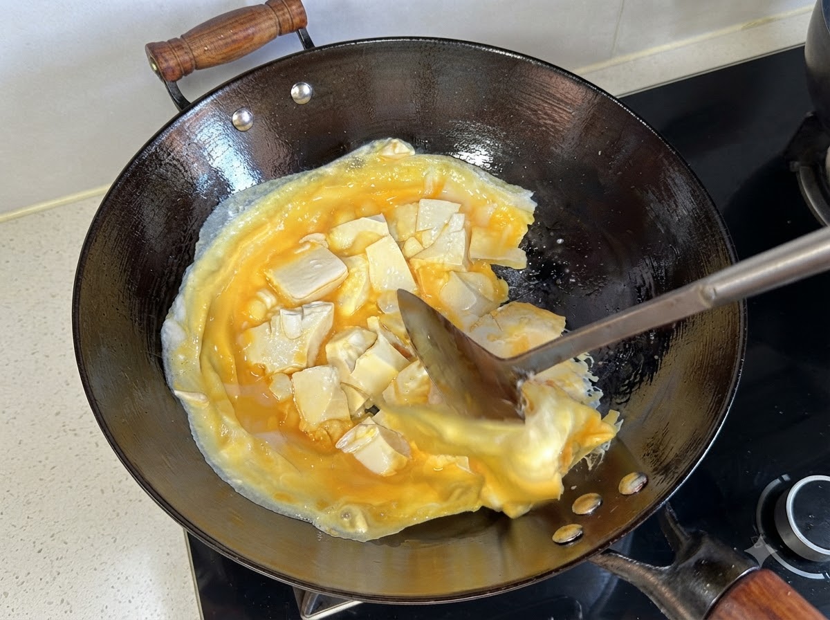 Una espátula levantando y dando la vuelta a la capa cocida de huevos y tofu en el wok.