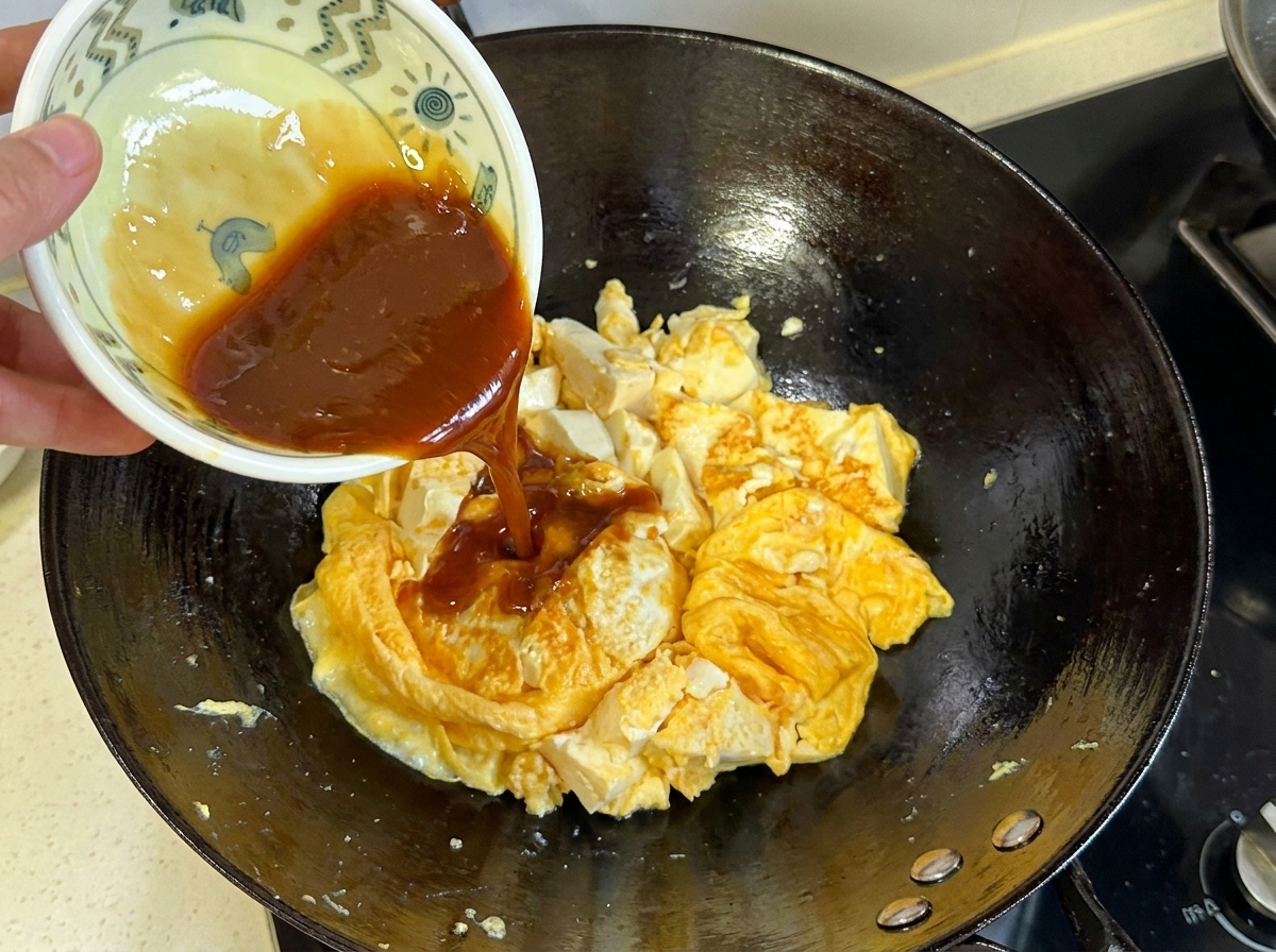 Salsa de color marrón oscuro vertiéndose desde un tazón pequeño sobre la mezcla cocida de huevo y tofu en el wok.