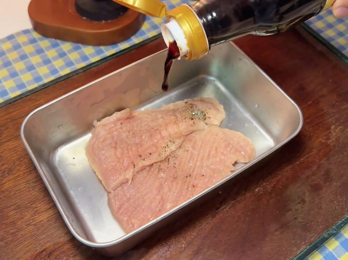 Salsa de soja oscura vertiéndose desde una botella de vidrio sobre chuletas de cerdo sazonadas en una bandeja de metal.