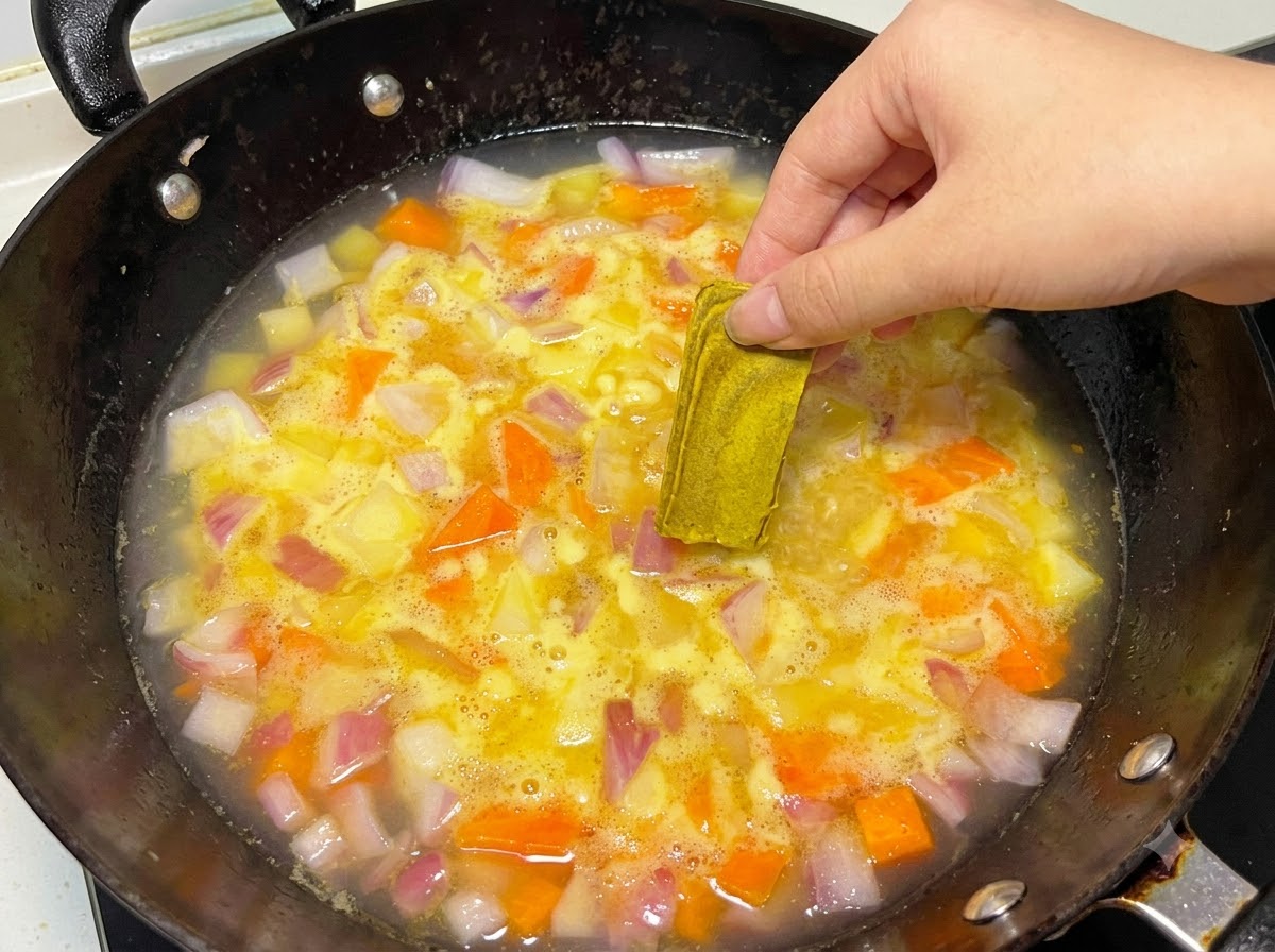 Una mano colocando un bloque sólido de curry en una olla hirviendo a fuego lento con agua y verduras.