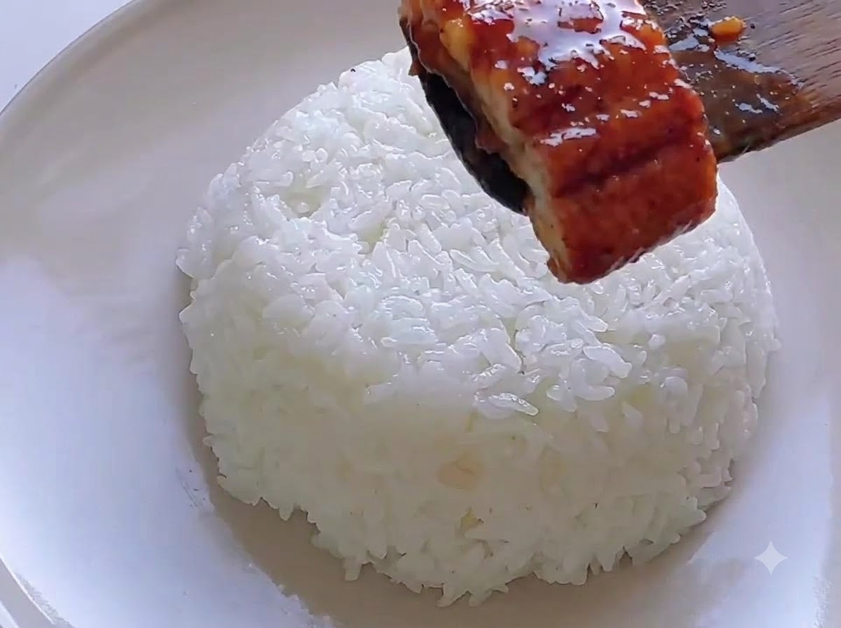 Una espátula de madera colocando un trozo de anguila glaseada sobre una porción de arroz blanco.