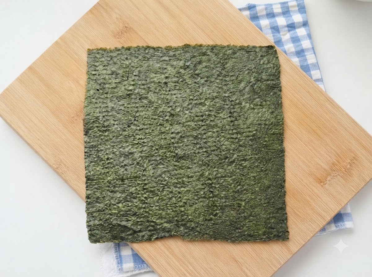 Una hoja entera de nori para sushi verde reposando sobre una tabla de cortar de madera con un paño de cuadros azules debajo.