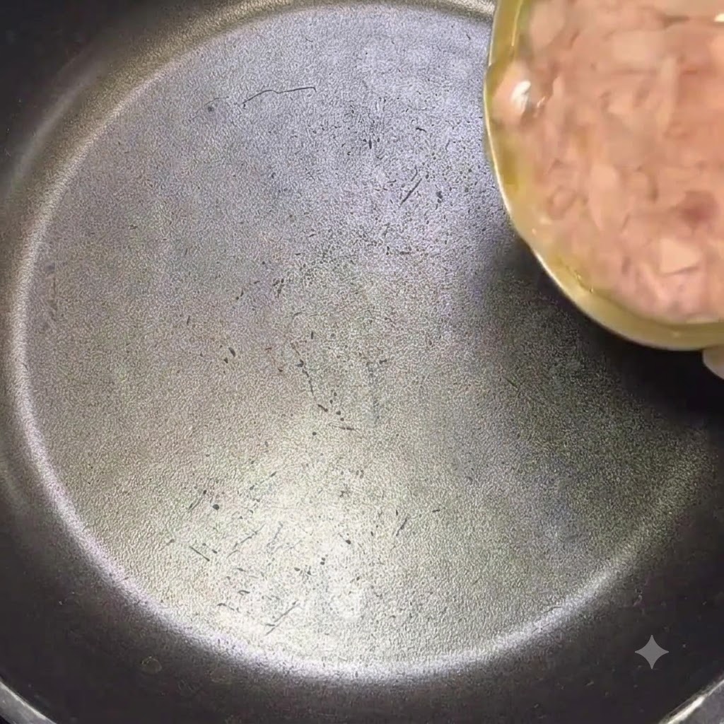 Una lata grande de atún siendo vertida con su aceite en una sartén negra.