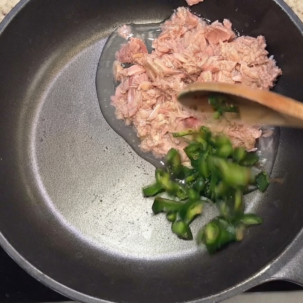 Rodajas de chiles verdes siendo añadidas a la sartén con el atún usando una cuchara de madera.