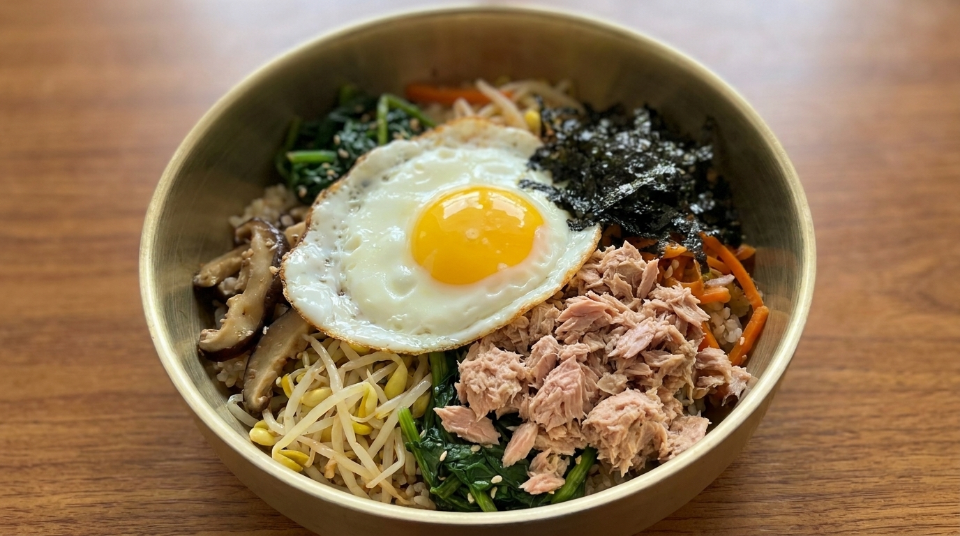 Un plato colorido de bibimbap de atún que incluye arroz sazonado, verduras frescas, setas, copos de alga, atún en lata y un huevo frito.