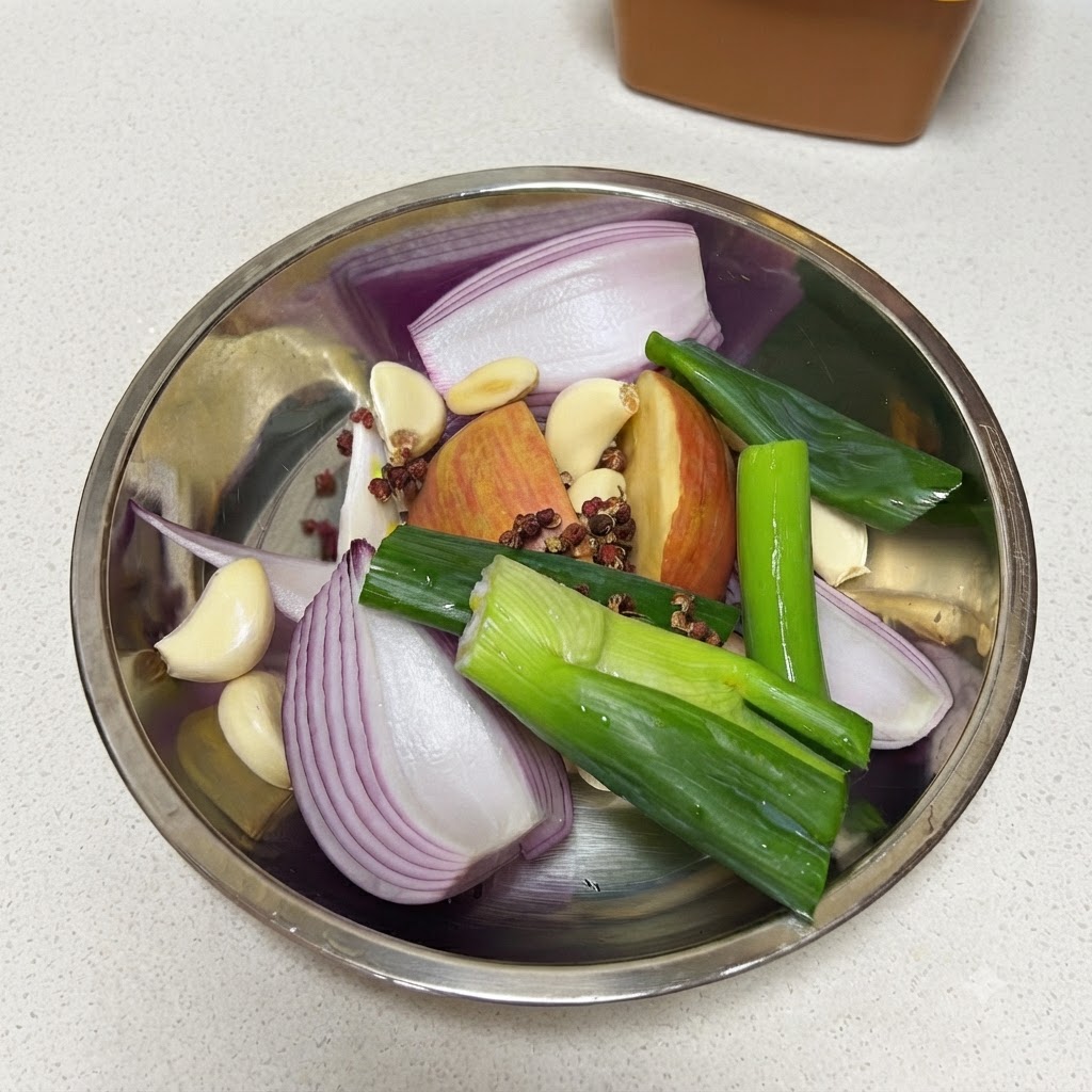 Cebollas verdes frescas siendo cortadas en un tazón de metal lleno de cebolla roja, manzana, dientes de ajo, granos de pimienta y una hoja de laurel.