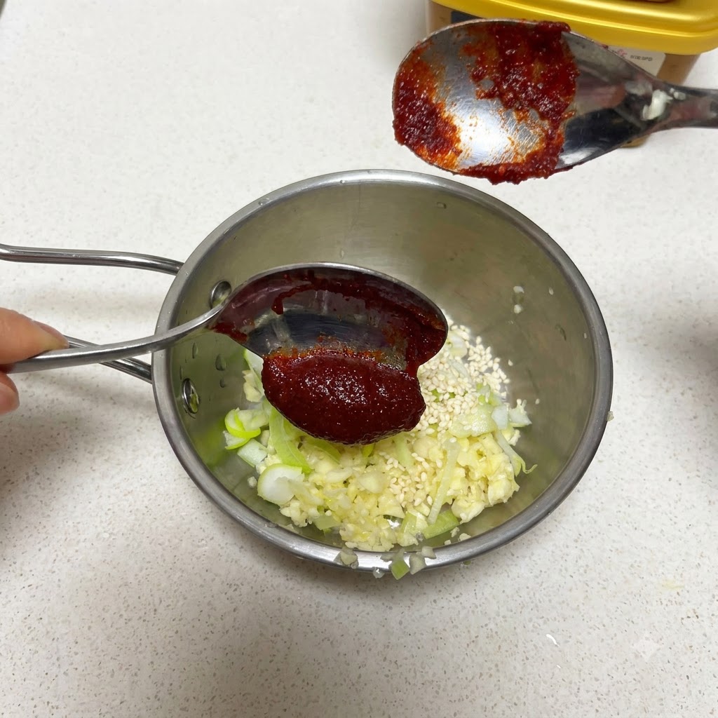 Una cucharada de pasta de chile coreana sostenida sobre una sartén con ajo y semillas de sésamo.