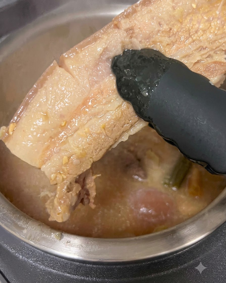 Un trozo grueso de panceta de cerdo cocida siendo levantado de una olla de caldo con pinzas negras.